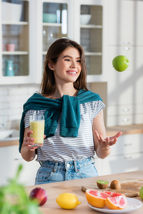 cheerful woman juggle with apple while holding fresh smoothie on blurred foreground