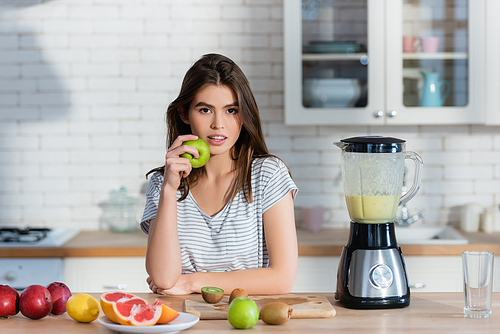 young woman  while holding apple near fresh fruits and blender
