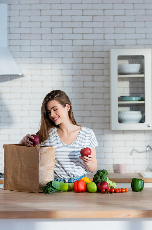 cheerful woman holding juicy apples near paper bag and fresh vegetables