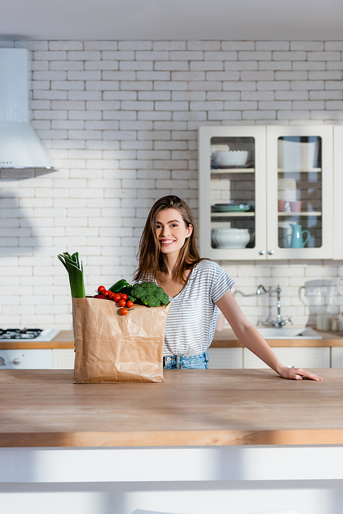 happy woman smiling at camera near paper bag with fresh vegetables in modern kitchen