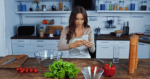 happy woman in apron holding smartphone near ingredients on table