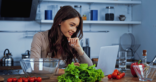 happy woman searching recipe in laptop near ingredients on table