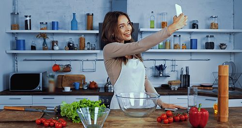 cheerful woman in apron taking selfie near ingredients on table
