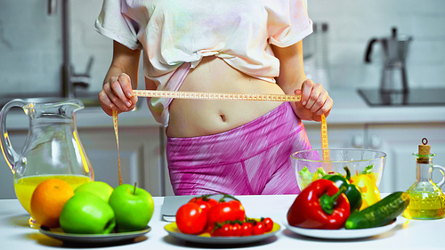 partial view of woman measuring waist near vegetables and fruits on blurred foreground