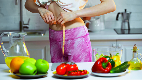 cropped view of woman measuring waist near vegetables and fruits on blurred foreground