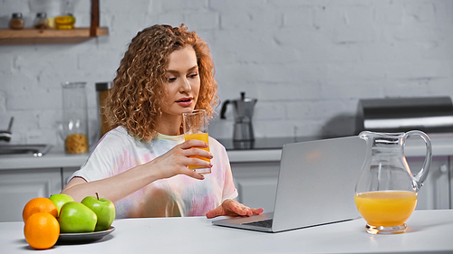 curly young woman looking at laptop while holding glass of orange juice near fruits on kitchen table