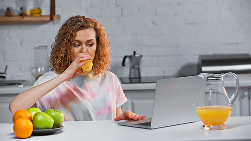 curly young woman looking at laptop while drinking orange juice
