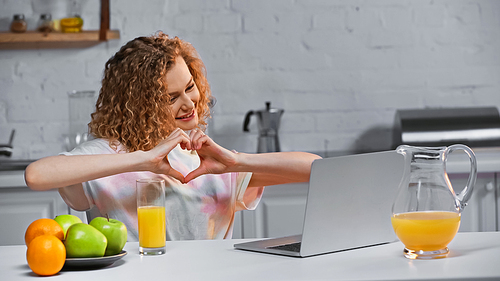 curly young woman looking at laptop while showing heart sign with hands during video call