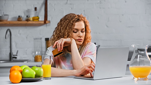 curly young woman holding credit card while shopping online in kitchen