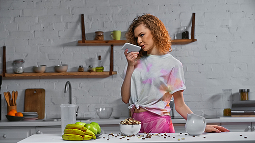 curly woman recording voice message near corn flakes and fruits on kitchen table