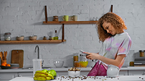 curly woman taking photo of corn flakes and fruits on kitchen table