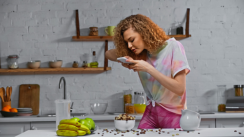 curly young woman taking photo of corn flakes and fruits on kitchen table