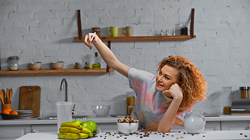 curly young woman taking selfie near corn flakes and fruits on kitchen table