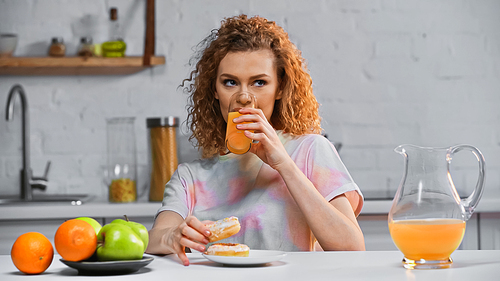 curly woman holding doughnut and drinking orange juice in kitchen