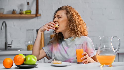 curly woman eating sweet doughnut and holding glass of orange juice in kitchen