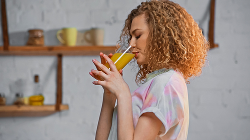 side view of curly woman drinking orange juice in kitchen