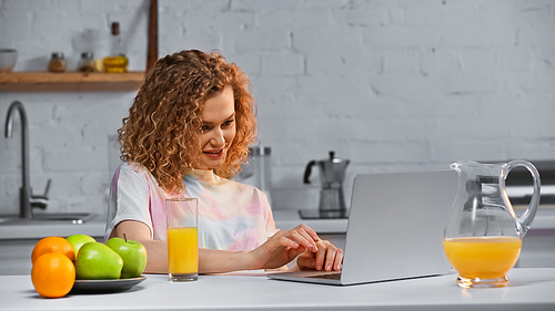 joyful woman looking at laptop near fruits and glass of orange juice on table