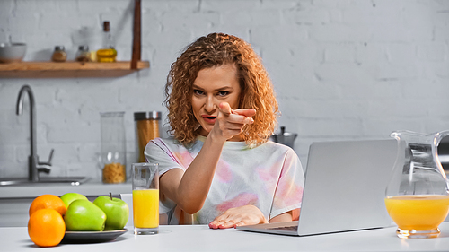 curly woman pointing with finger while  near laptop and fresh fruits on table