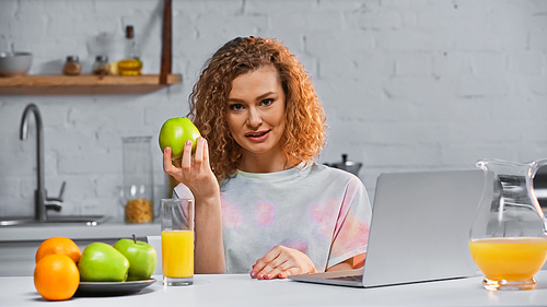 curly woman holding apple while  near laptop and fruits on table