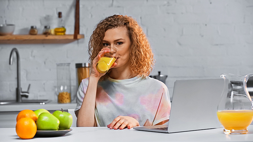 curly woman drinking orange juice while  near laptop and fruits on table