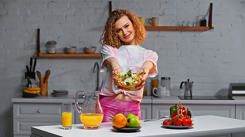 smiling and curly woman holding bowl with salad in kitchen