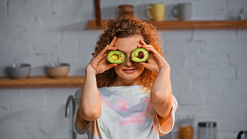 cheerful young woman covering eyes with halves of avocado