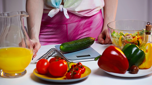 cropped view of woman writing in notebook while weighing cucumber
