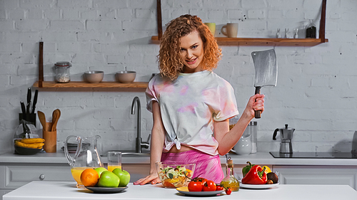cheerful woman holding huge knife near fresh salad in bowl and fruits on table