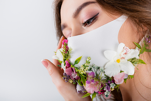 Portrait of young woman holding hand near medical mask with flowers isolated on grey