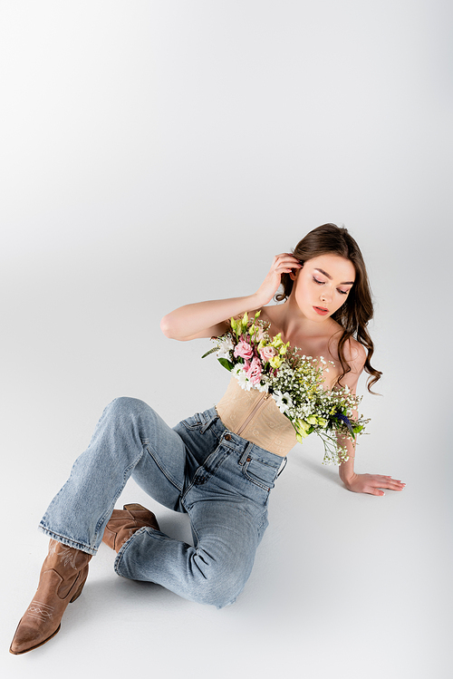 Brunette woman in blouse with flowers and jeans sitting on grey background