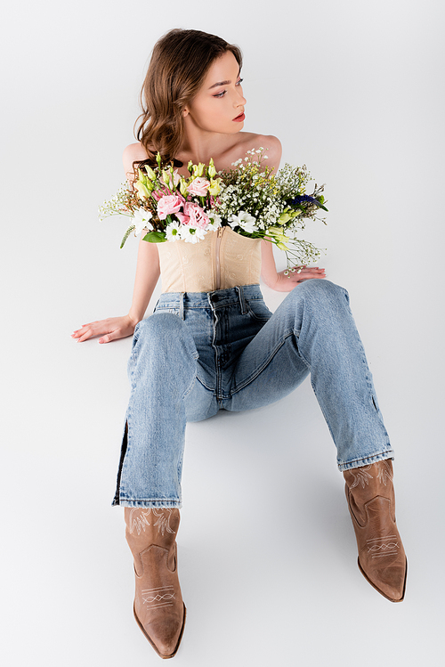 Stylish woman in brown shoes, jeans and blouse with different flowers sitting on grey background