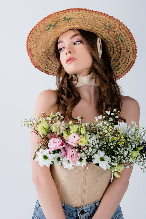 Curly model in sun hat with flowers in blouse looking away isolated on grey