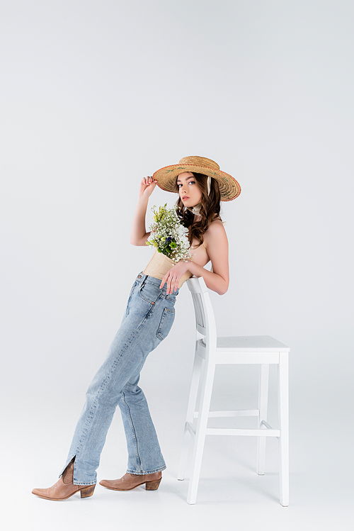 Trendy model with flowers in blouse standing near chair on grey background