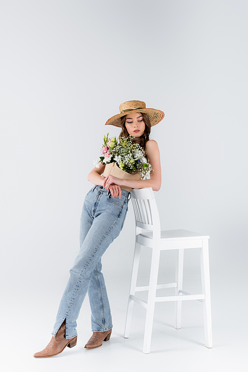 Young model in jeans, shoes and flowers in blouse standing near chair on grey background