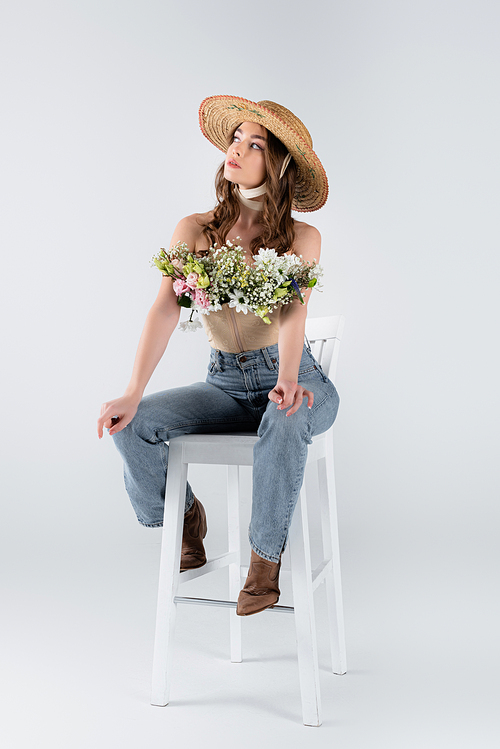 Young woman in sun hat and flowers in blouse looking away on chair on grey background
