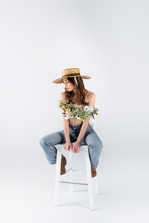 Trendy woman in straw hat and floral bouquet in blouse posing on chair on grey background