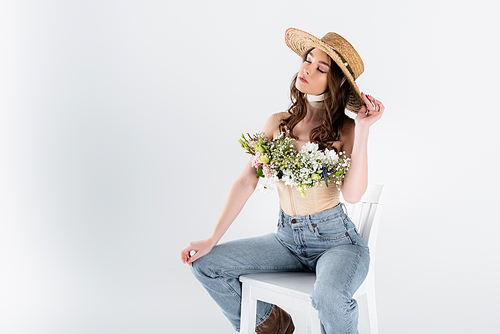 Young woman with flowers and sun hat sitting on white chair isolated on grey