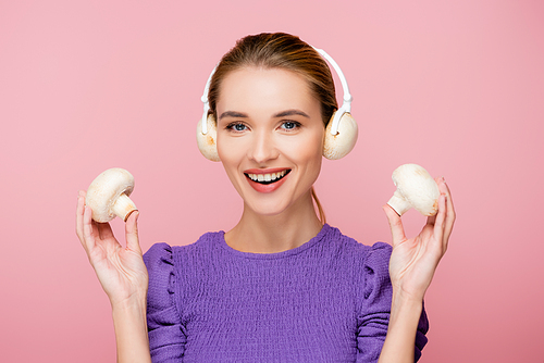 smiling woman with mushrooms in headphones holding champignons isolated on pink
