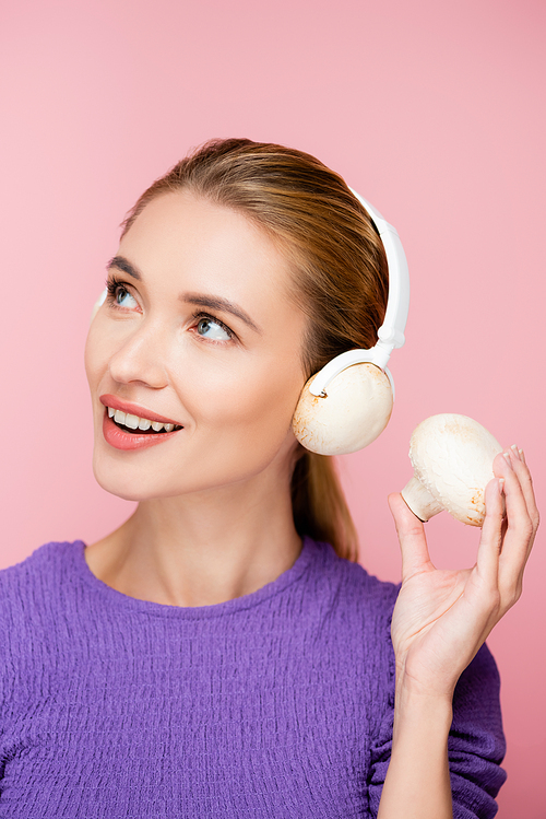 cheerful woman with mushroom in headphones looking away isolated on pink