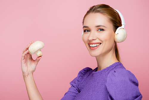 young woman with mushroom in headphones smiling at camera isolated on pink