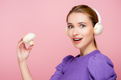 excited woman with mushrooms in headphones holding champignon isolated on pink