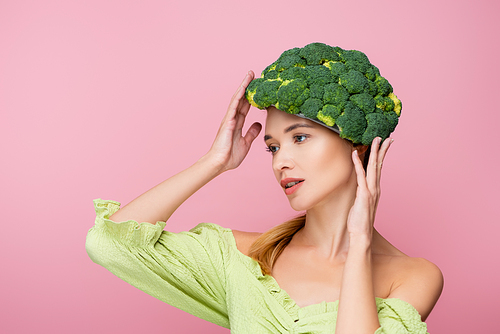 young woman adjusting hat made of broccoli while posing isolated on pink, surrealism concept