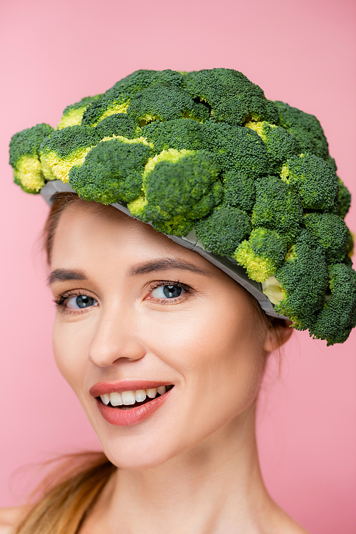 portrait of cheerful woman in hat made of fresh broccoli isolated on pink, surrealism concept