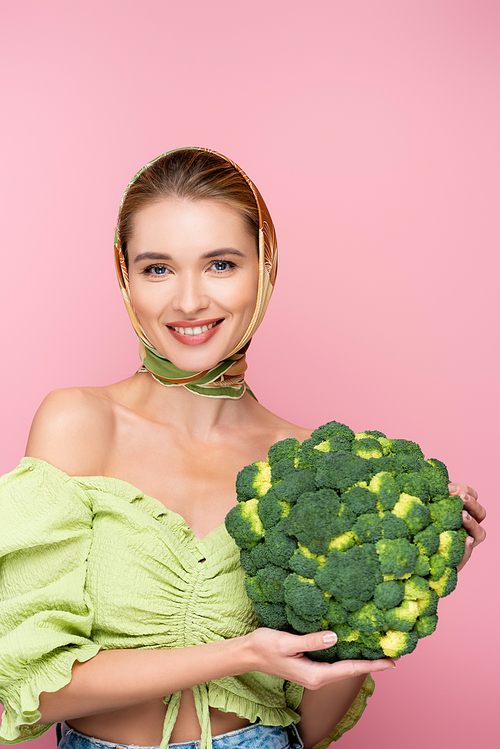 happy woman in kerchief holding green broccoli while smiling at camera isolated on pink, surrealism concept