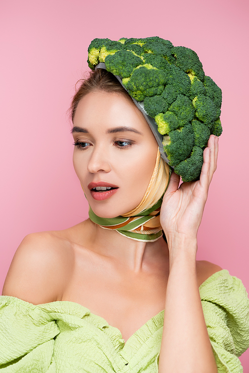 charming woman touching hat made of green fresh broccoli isolated on pink, surrealism concept