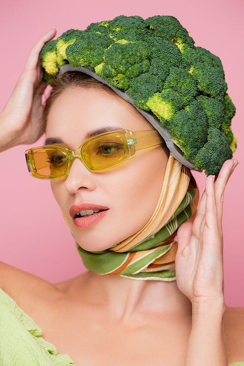 young woman in colored eyeglasses touching hat made of fresh broccoli isolated on pink, surrealism concept