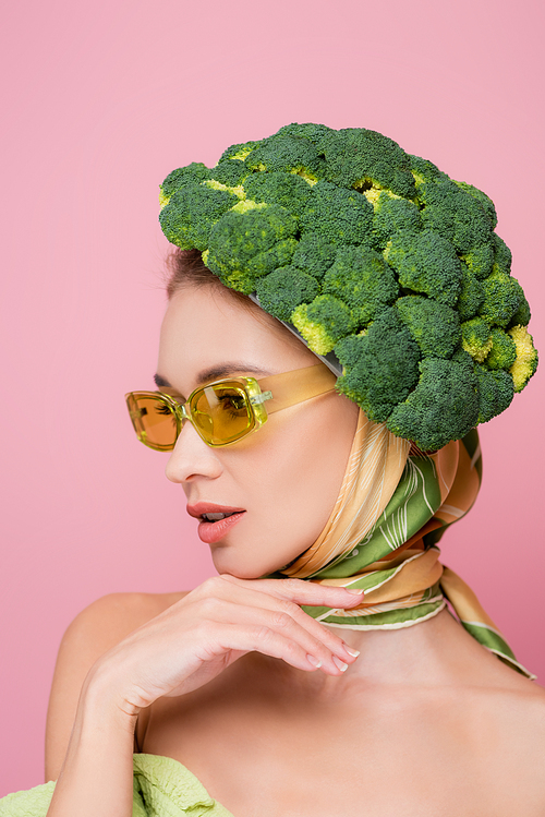 charming woman in colored eyeglasses, posing in hat made of broccoli isolated on pink, surrealism concept