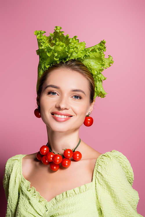 happy woman in necklace, earrings and hat made of vegetables smiling at camera on pink