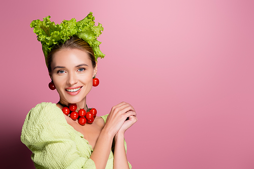smiling woman in hat, necklace and earrings made of vegetables posing on pink