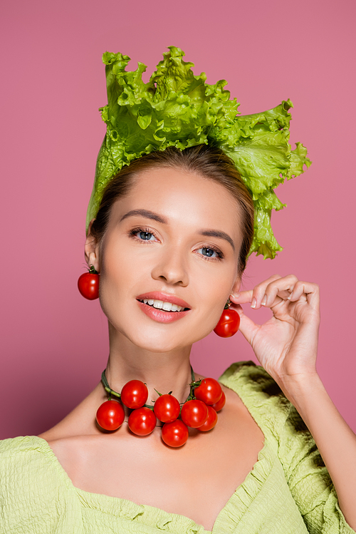 smiling woman in hat, necklace and earrings made of fresh vegetables posing on pink